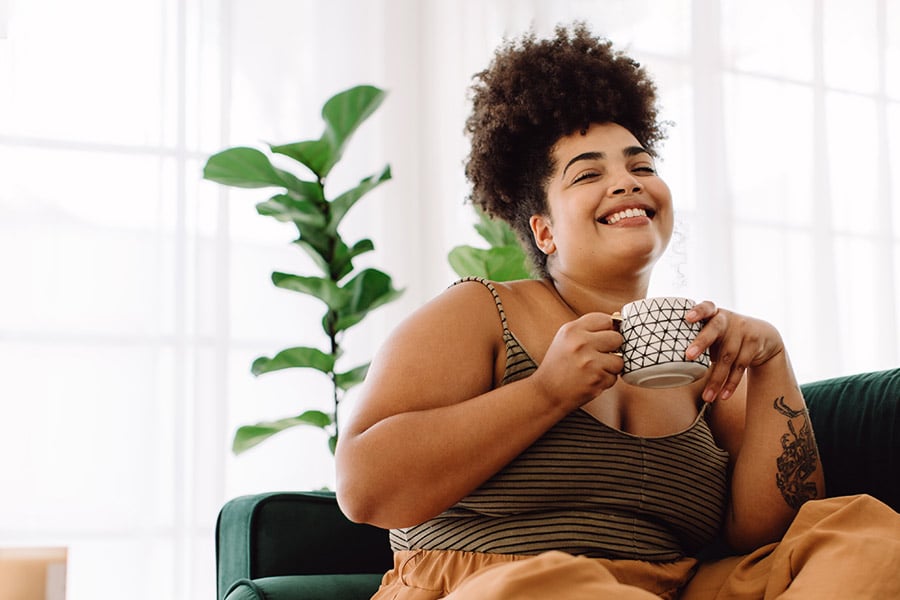 Smiling Woman Drinking Coffee on a Green Couch With House Plants Behind Her